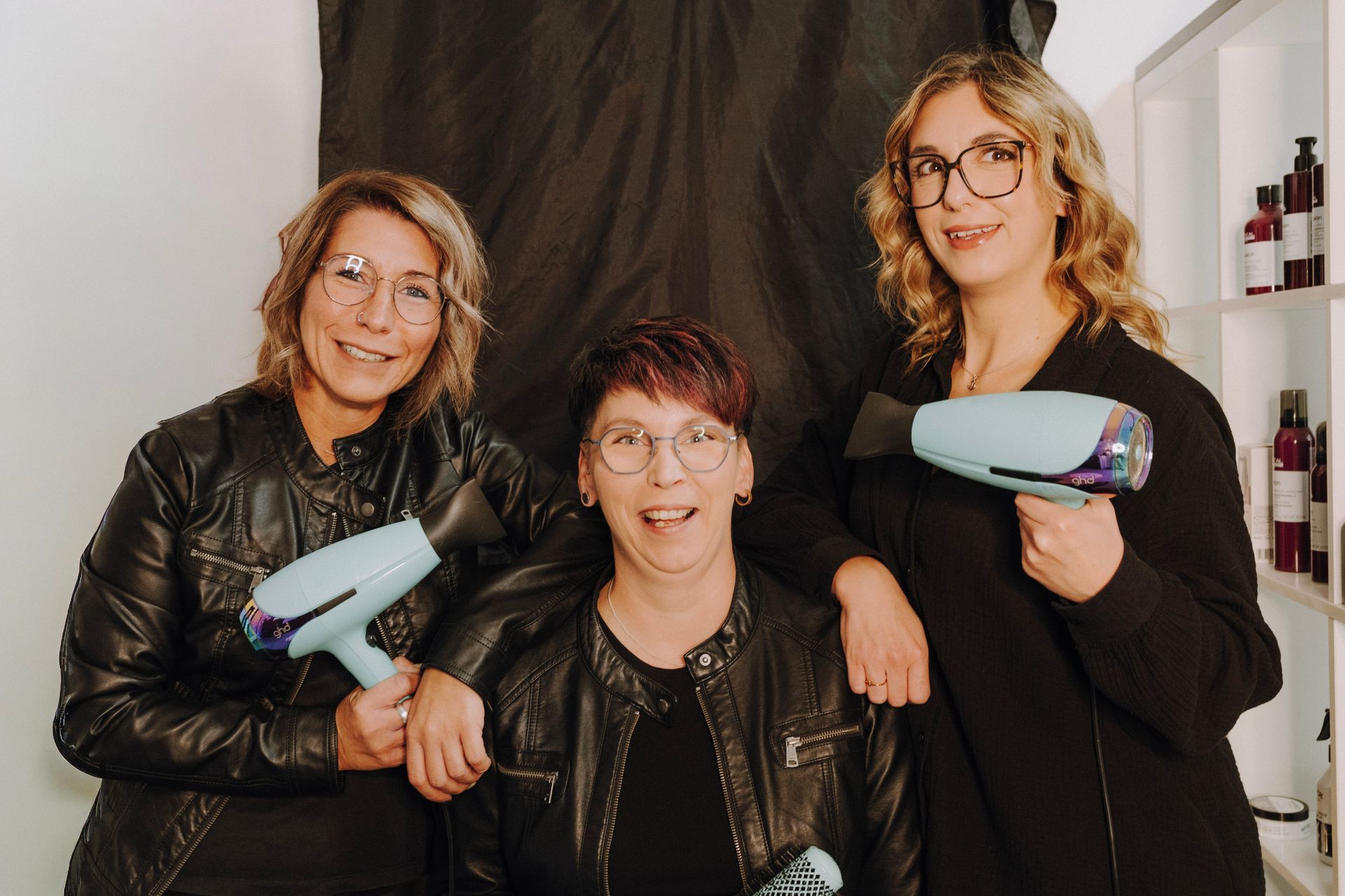 Drei Personen mit Haartrocknern posieren in einem Friseursalon. Zwei Frauen in Lederjacken lehnen sich in der Mitte aneinander.