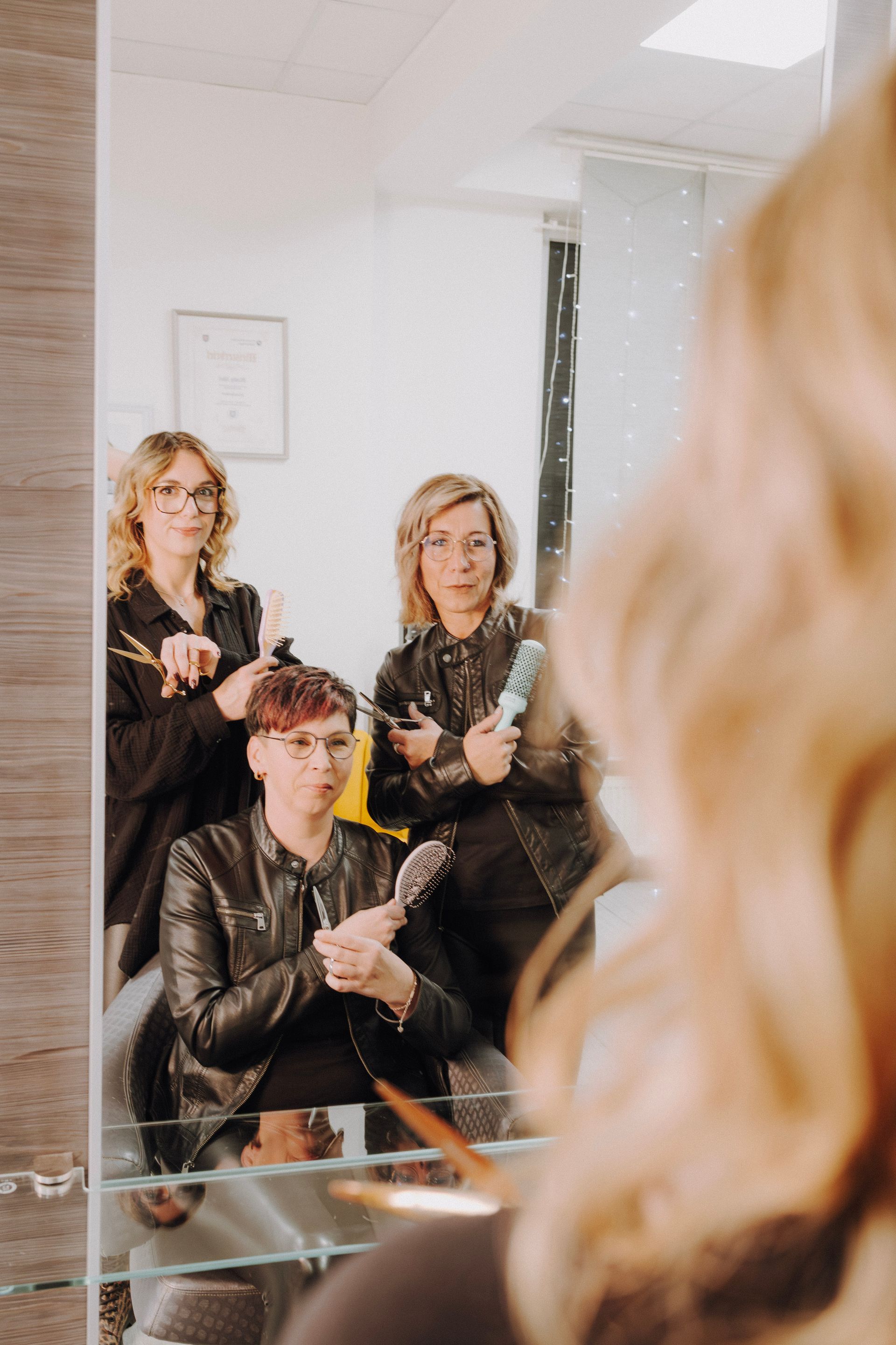 Drei Friseure mit Werkzeugen, deren Spiegelbild zu sehen ist. Eine Person im Vordergrund. Alle tragen schwarze Jacken.