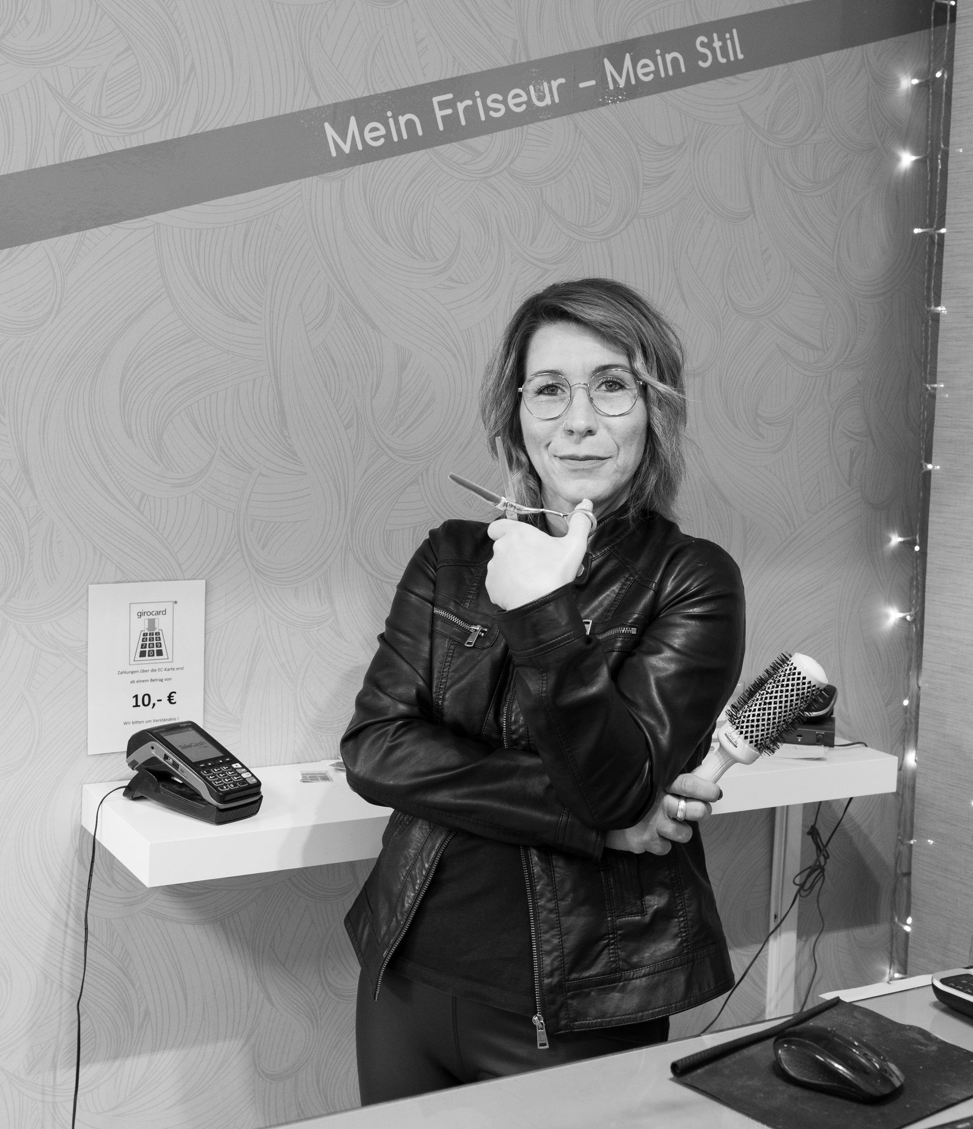 Frau mit Brille und schwarzer Lederjacke, die in einem Friseursalon eine Schere und eine Haarbürste hält.