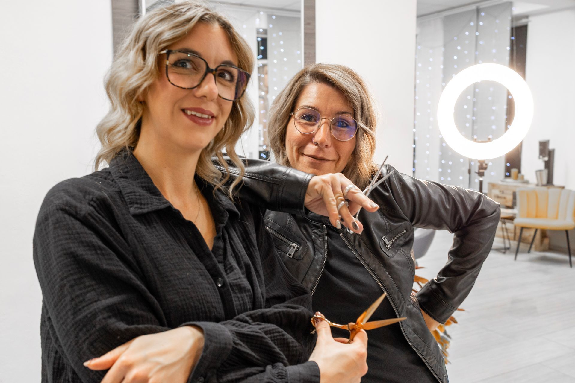 Zwei Frauen in einem Friseursalon; die eine hält eine Schere und deutet damit auf die andere, beide tragen Brillen und lächeln.