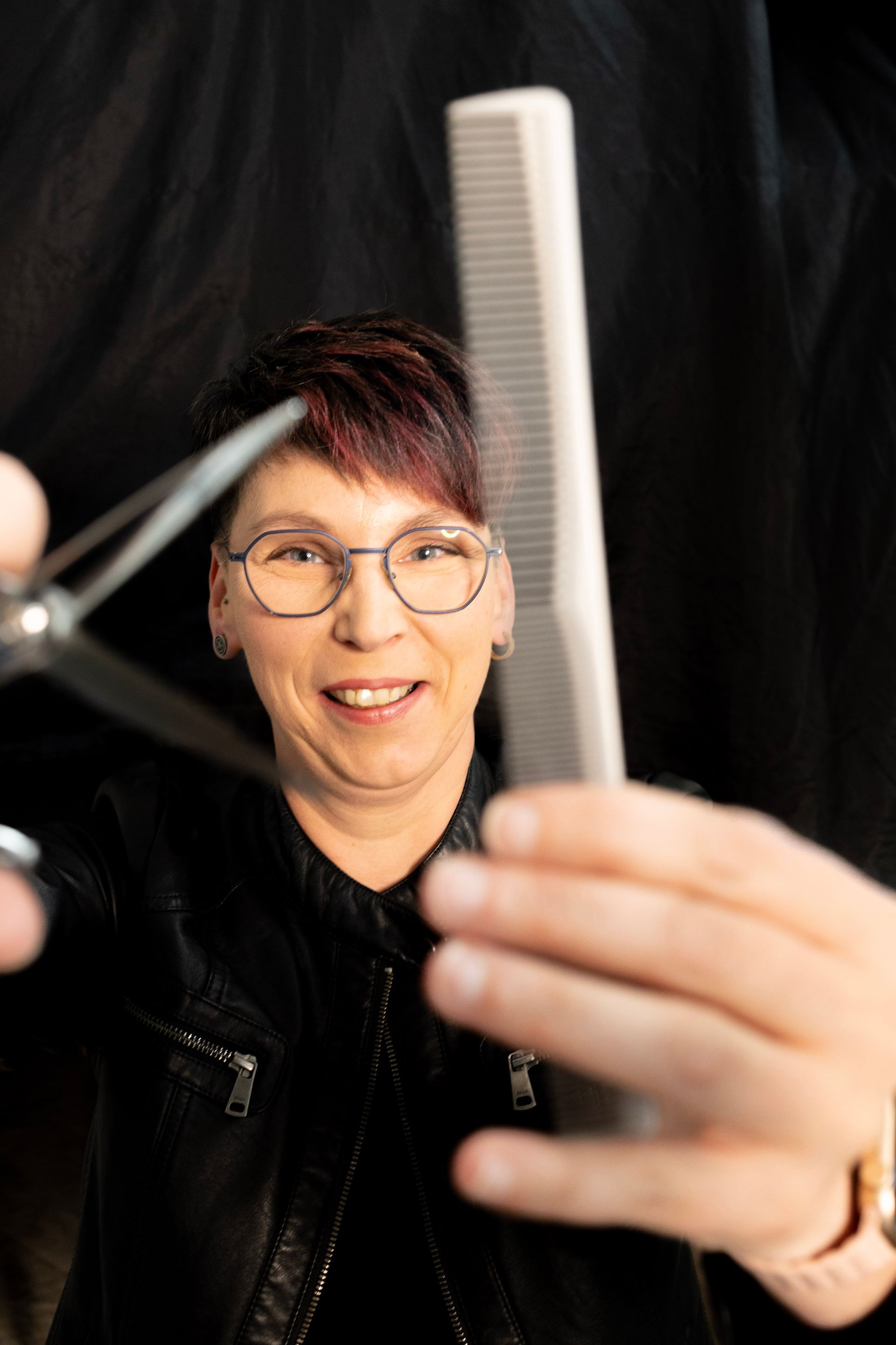 Frau hält Schere und Kamm und lächelt vor schwarzem Hintergrund in die Kamera.