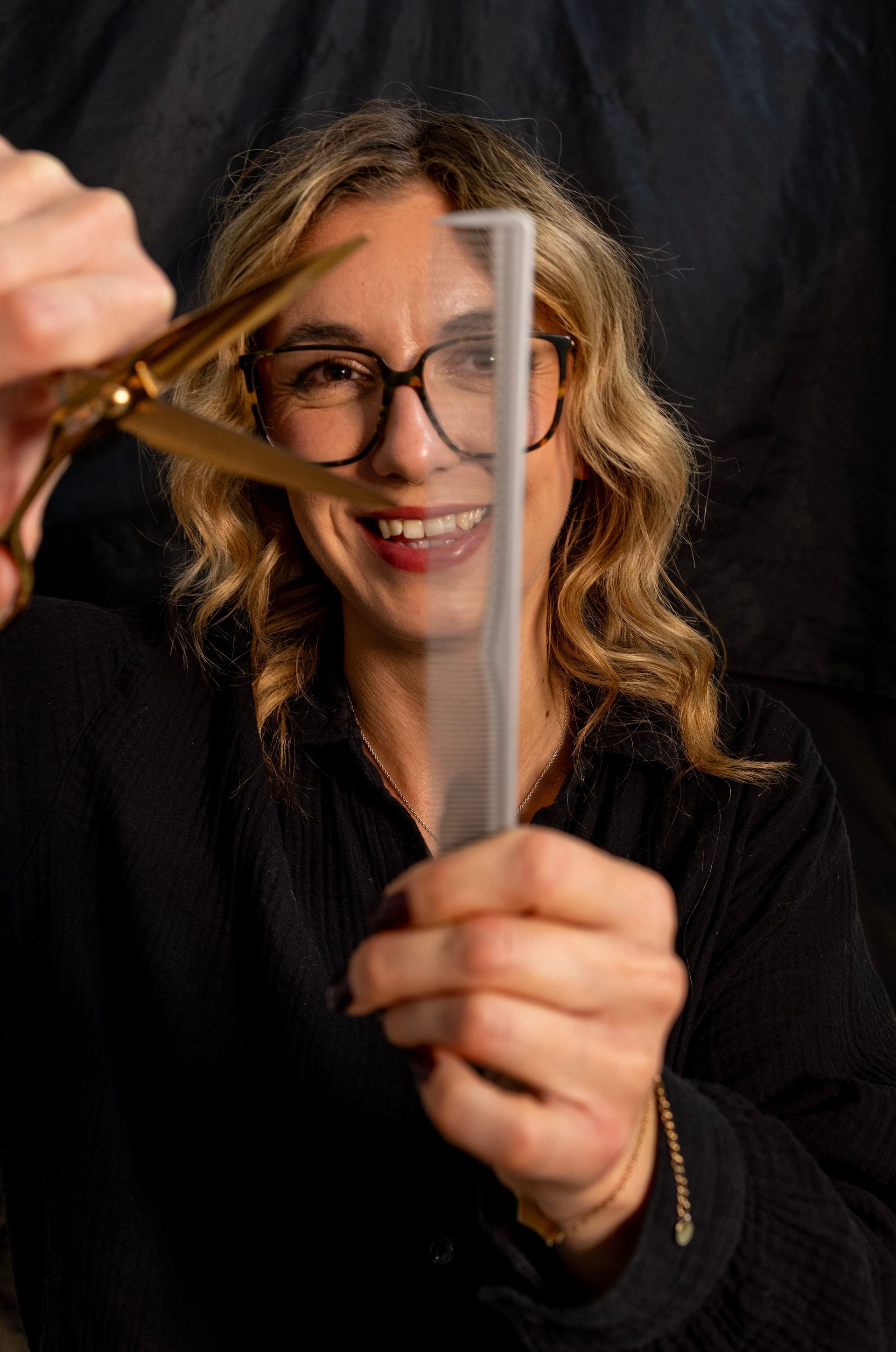 Frau mit Brille lächelt, hält Schere und Kamm. Dunkler Hintergrund.