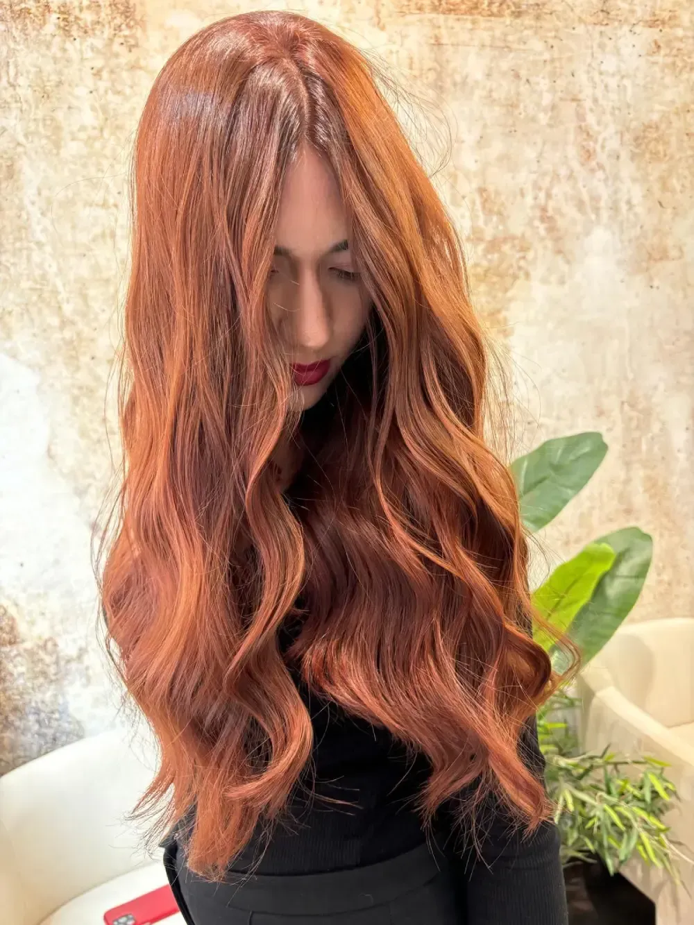 Mujer con cabello castaño rojizo largo y ondulado. De pie en un interior cerca de una planta.