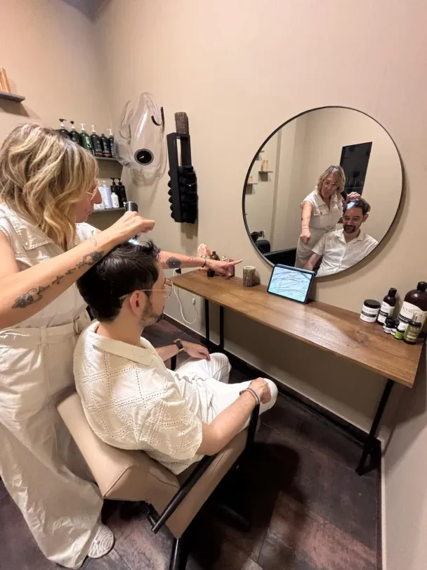 Un estilista peina a una clienta en un salón. La clienta mira su tableta mientras otro estilista le da consejos en el espejo.