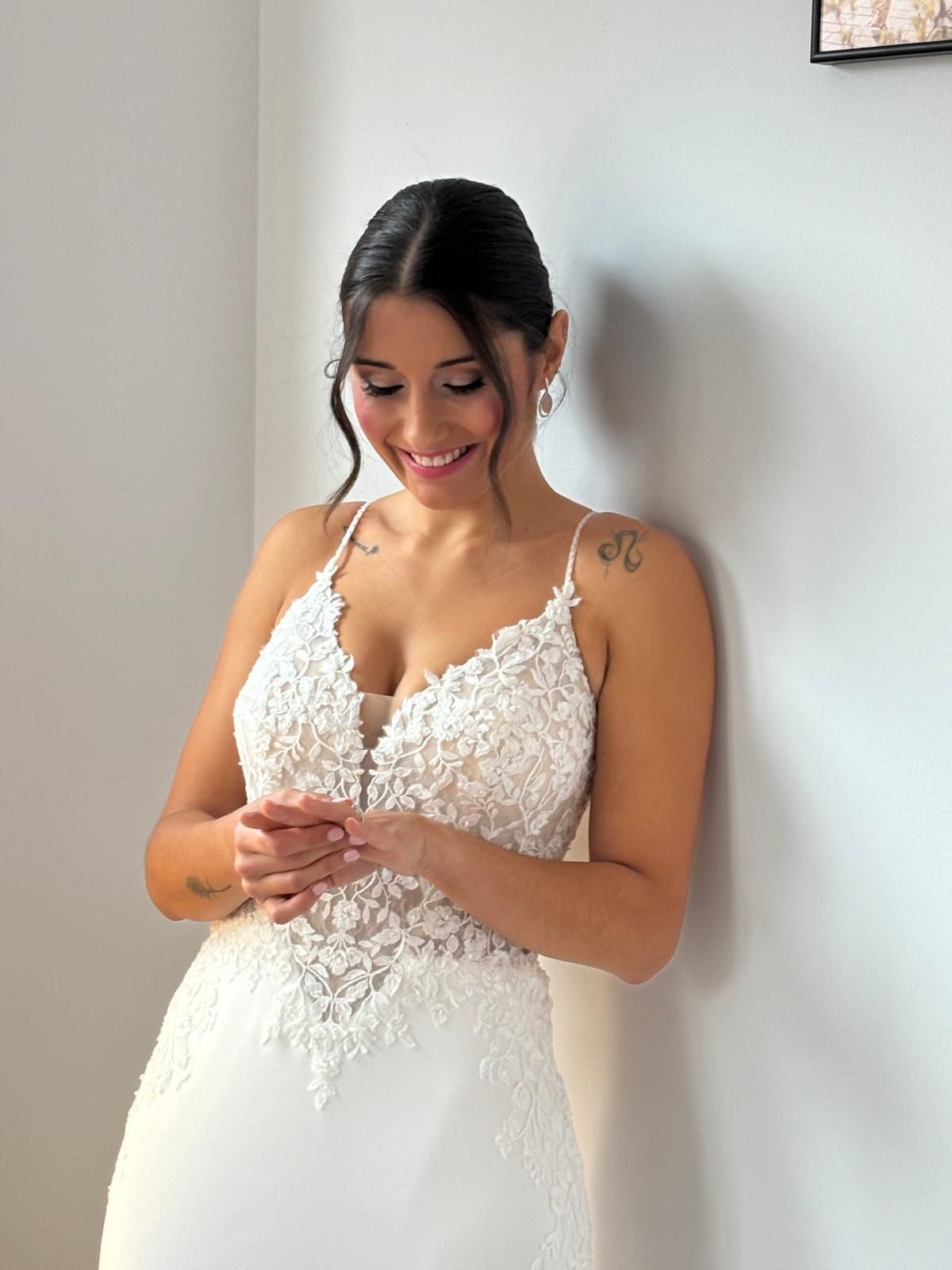 Una mujer con un vestido de novia de encaje blanco sonríe, apoyada contra una pared blanca.