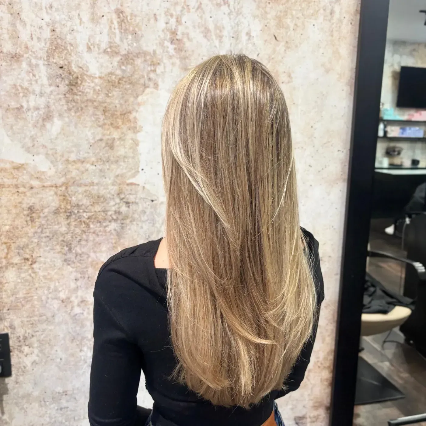 Una mujer con cabello rubio largo y en capas, vista desde atrás, se encuentra en un salón.