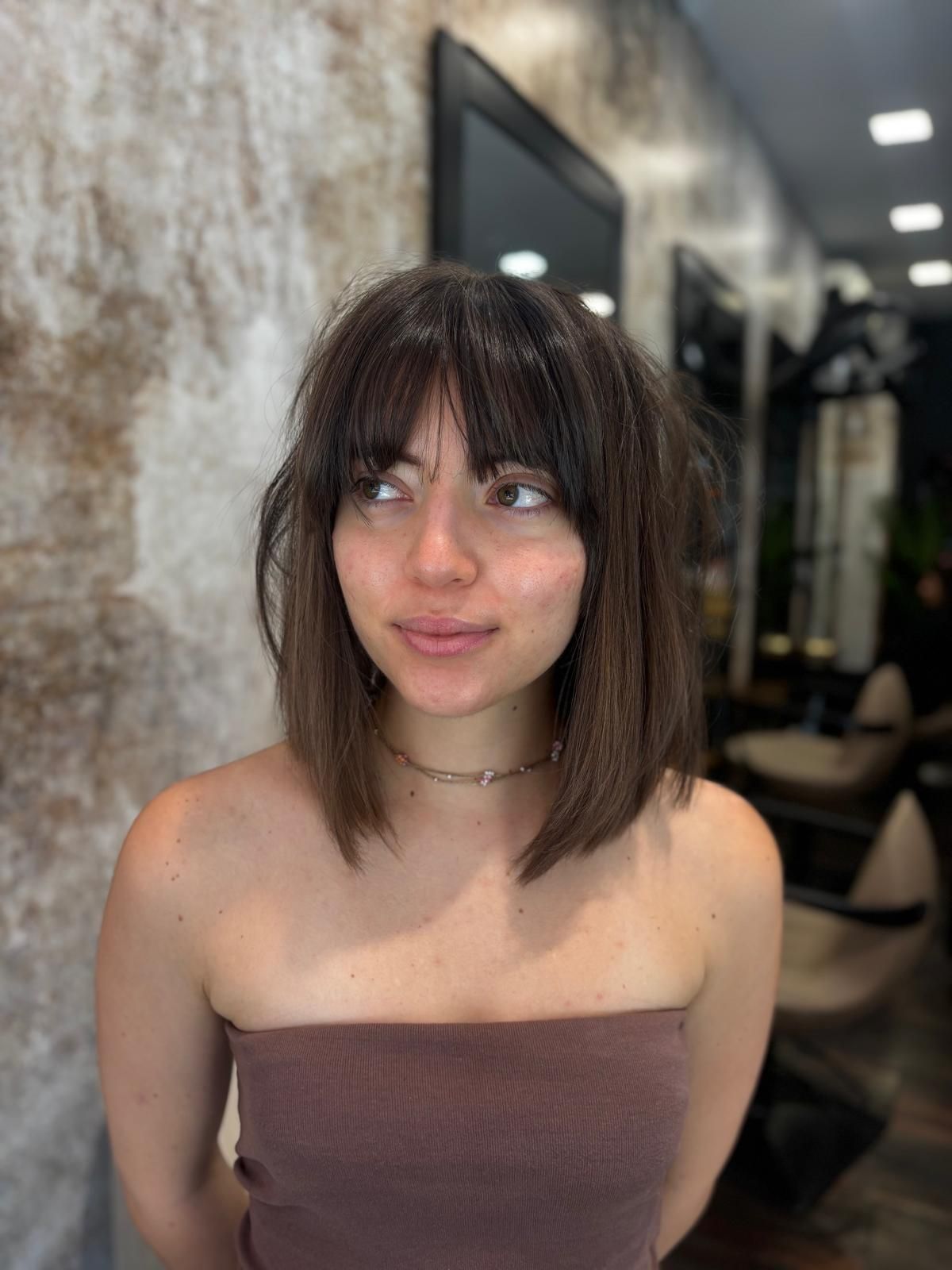 Mujer con cabello castaño hasta los hombros y flequillo con un top marrón sin tirantes en un salón.