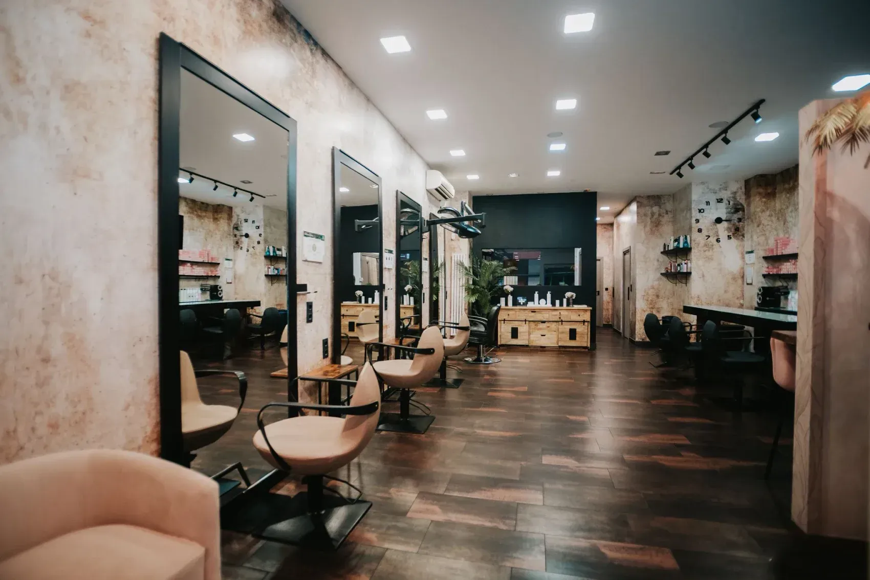 Interior de peluquería: sala larga con espejos, sillas de peluquería, suelo de madera y pared decorativa.