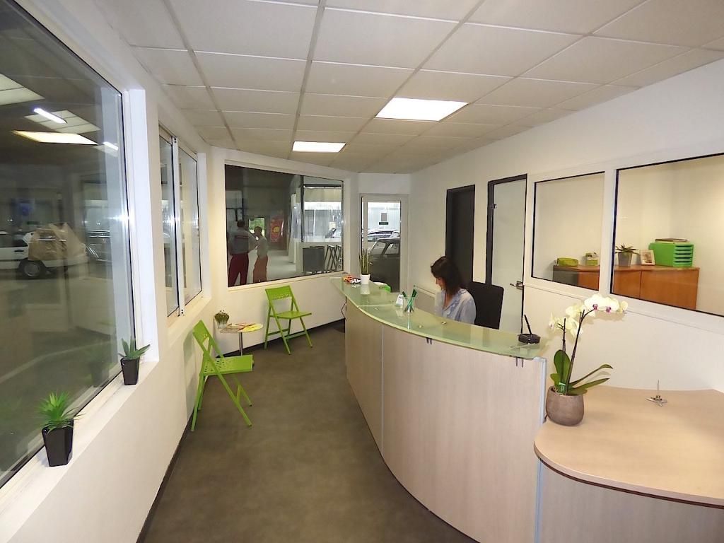 Espace de réception avec un bureau incurvé et une personne assise derrière. Chaises vertes et fenêtres présentes.