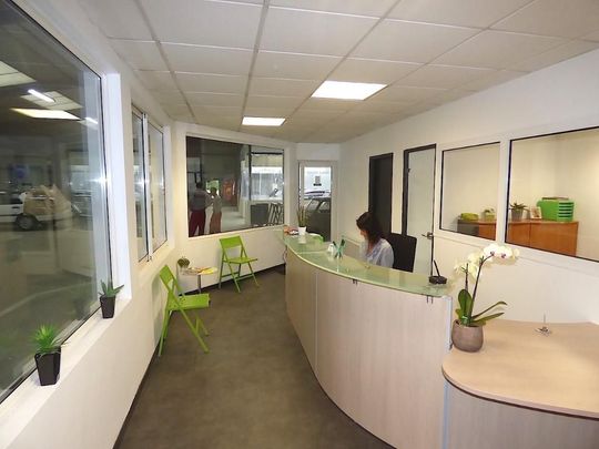 Espace de réception avec un bureau incurvé et une personne assise derrière. Des chaises vertes et des fenêtres sont visibles.