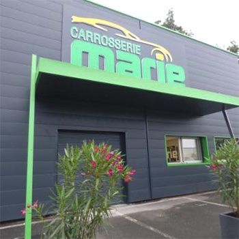 Extérieur de l'atelier de la Carrosserie Marie, bâtiment gris avec des accents verts, enseigne, plantes.