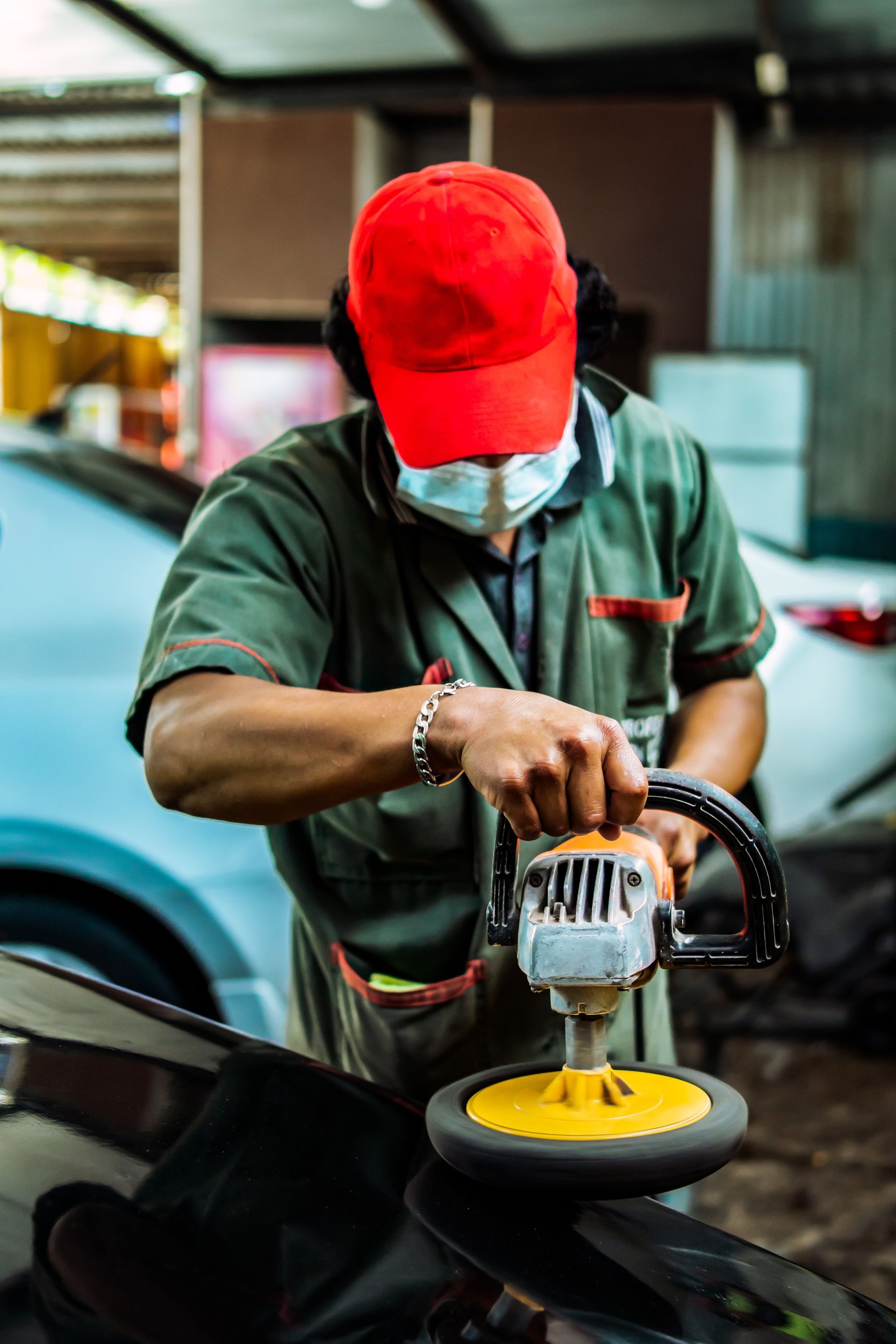 Une personne portant une casquette rouge et un masque polit une voiture avec une machine.