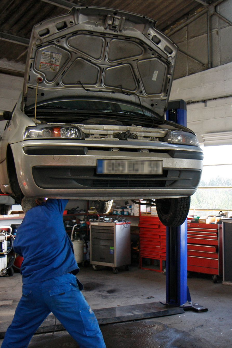 Un mécanicien en salopette bleue travaille sous une voiture argentée soulevée par un pont élévateur dans un garage.