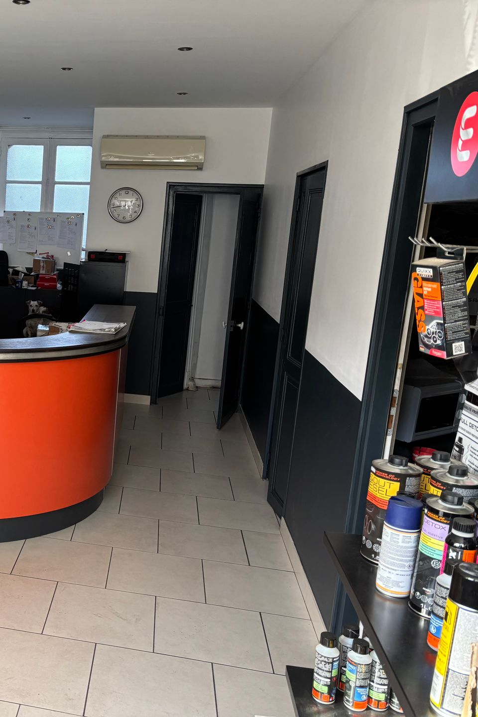 Vue intérieure d'un magasin avec un comptoir orange, des produits sur des étagères et des portes le long d'un couloir.