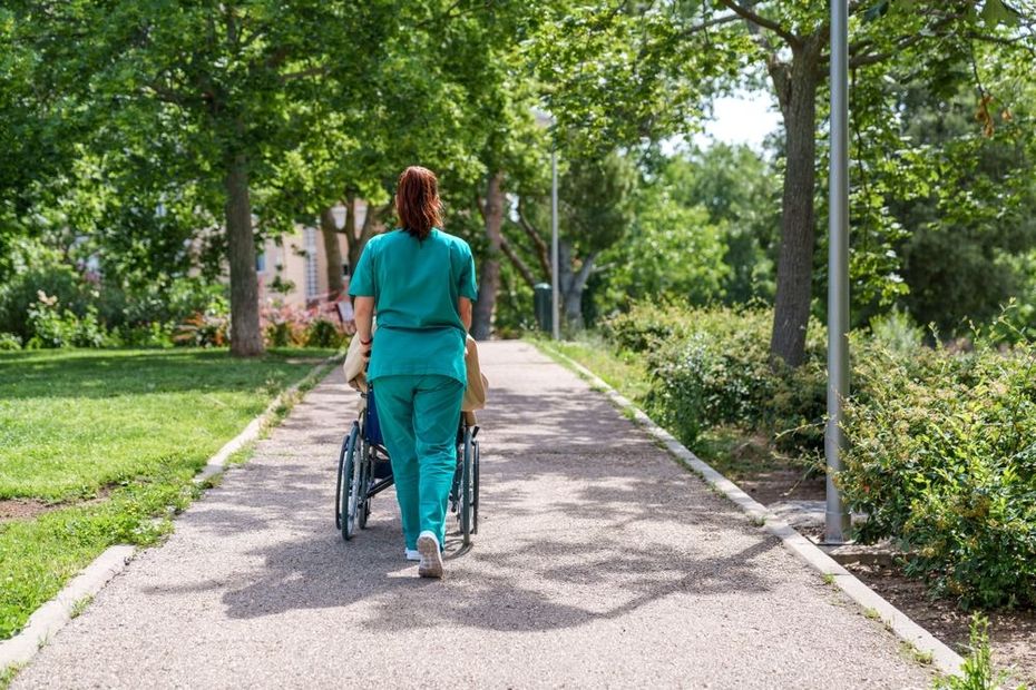 Una persona con uniforme quirúrgico color turquesa empuja una silla de ruedas por un sendero arbolado en un parque.