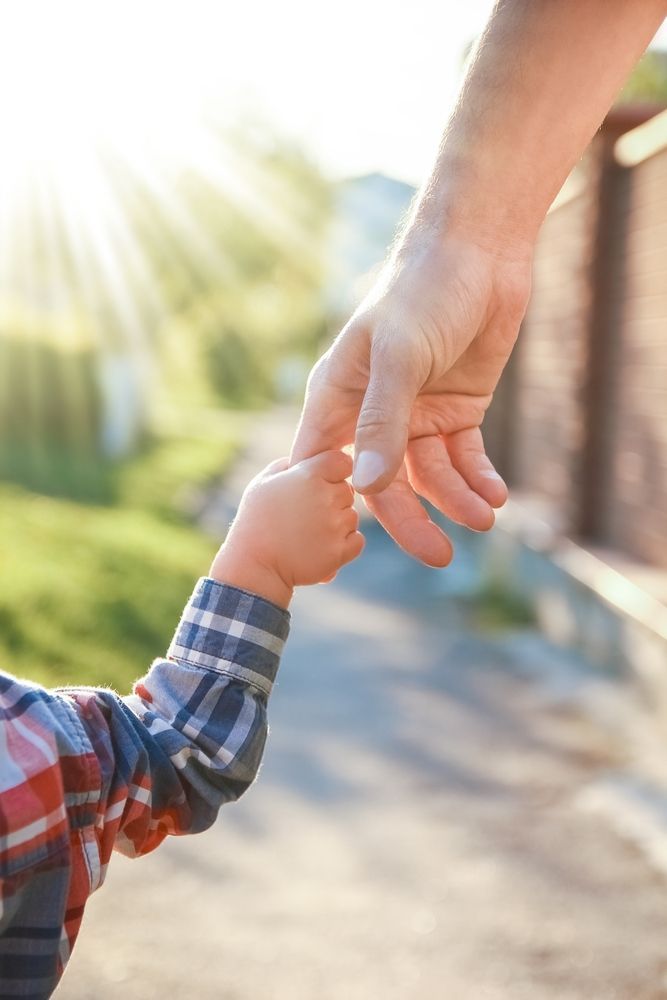 Una mano adulta sujeta la mano de un niño al aire libre, bajo la luz del sol, con un camino borroso y vegetación.