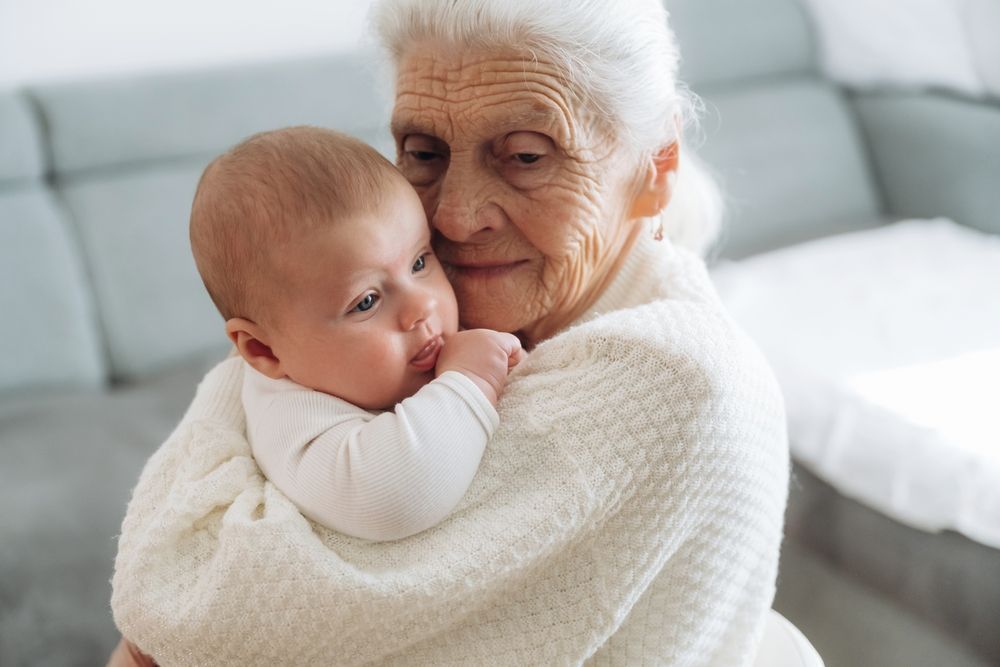 Una anciana con un suéter blanco sostiene con ternura a un bebé en un sofá.