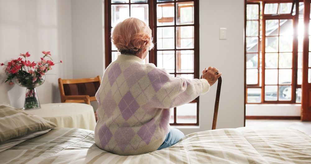 Una persona sentada en una cama estirándose en un dormitorio luminoso con ventanas de madera y un jarrón de flores.