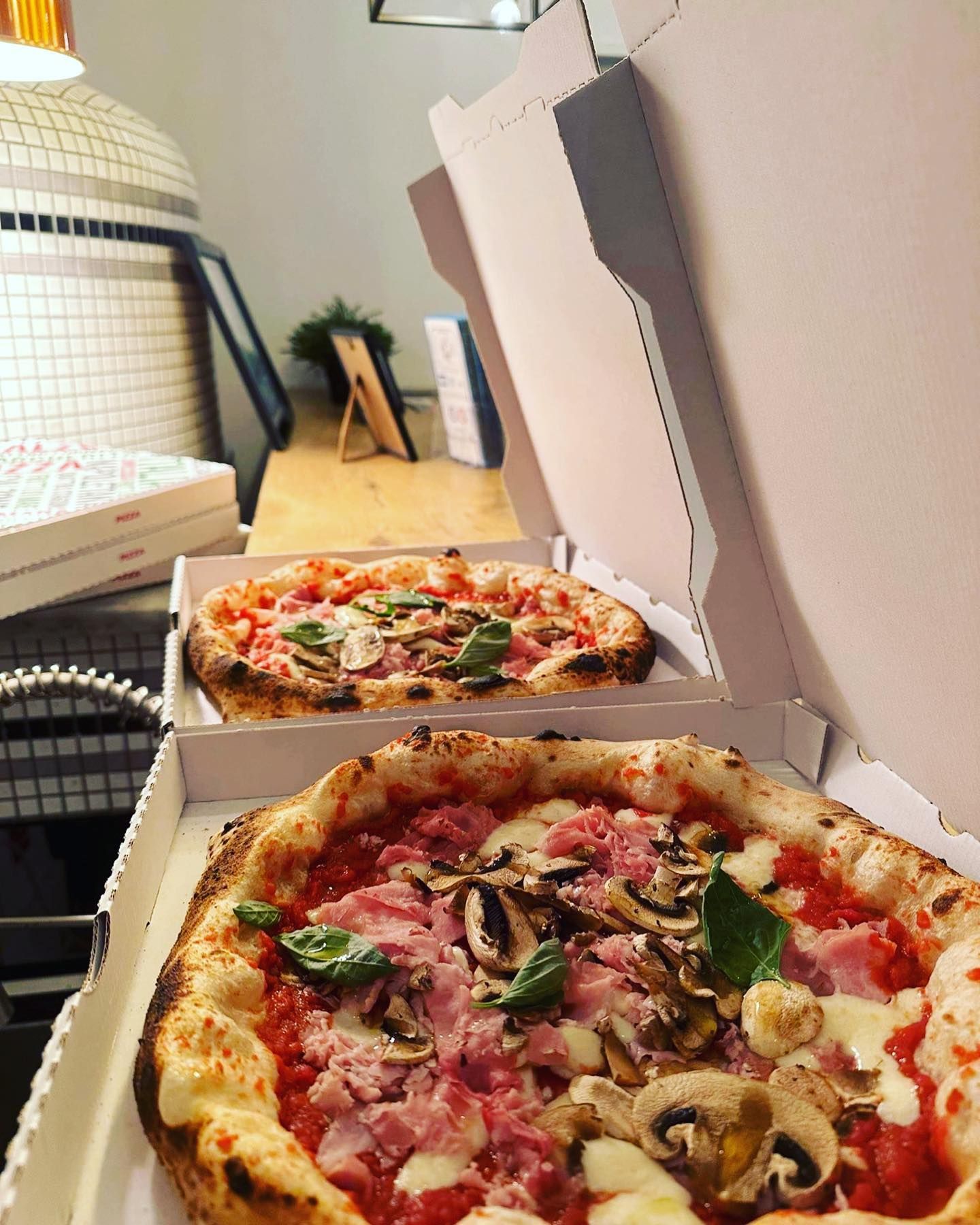 Deux pizzas en boîte, prêtes à être livrées. L'une est garnie de jambon et de champignons. On aperçoit le four en arrière-plan.