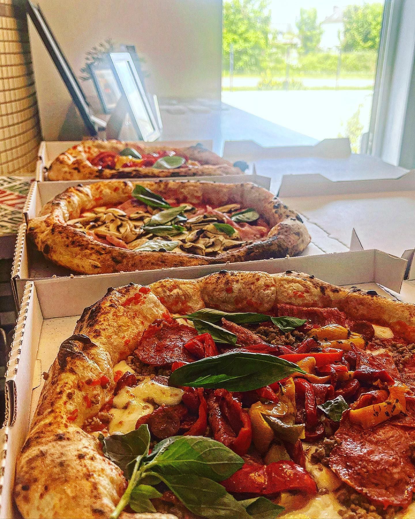 Trois pizzas dans des boîtes en carton ouvertes, posées sur un comptoir. Garnitures variées, dont basilic et pepperoni.