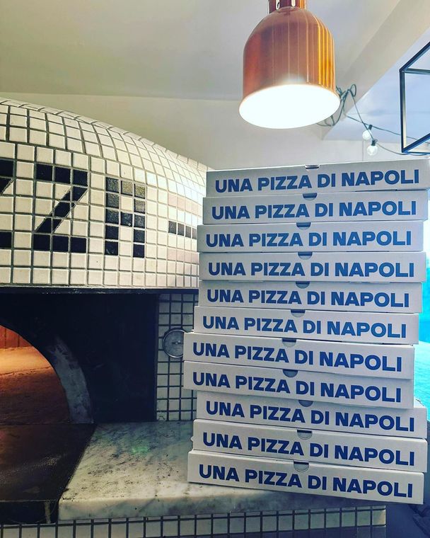 Pile de boîtes à pizza portant l'inscription Una Pizza Di Napoli à côté d'un four à pizza.