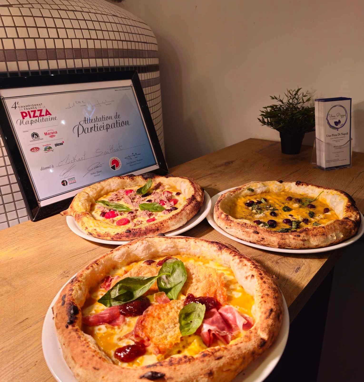 Trois pizzas gastronomiques sur une table, avec un certificat et un four en arrière-plan.
