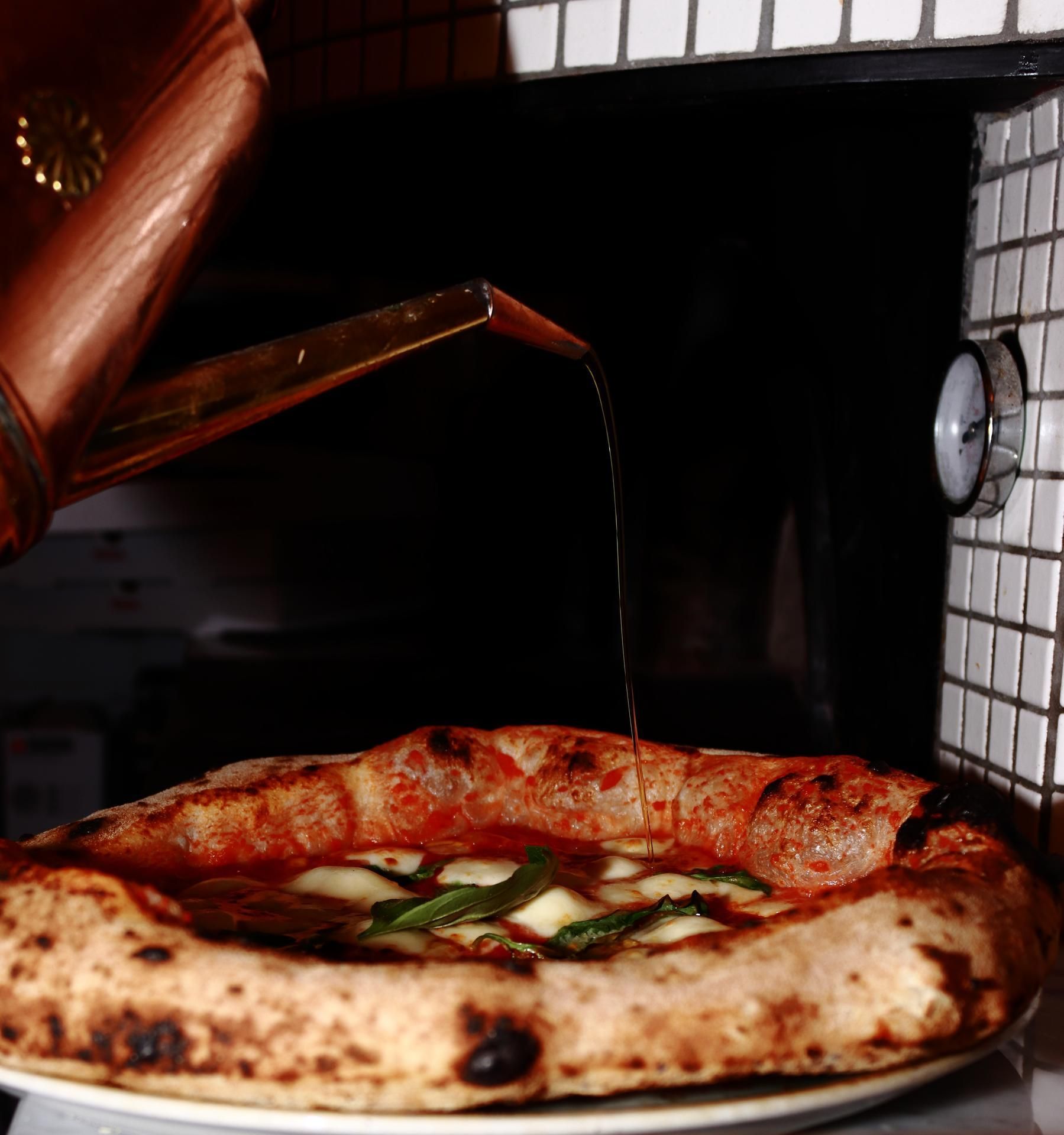 De l'huile d'olive est versée sur une pizza Margherita, près d'un four à pizza.
