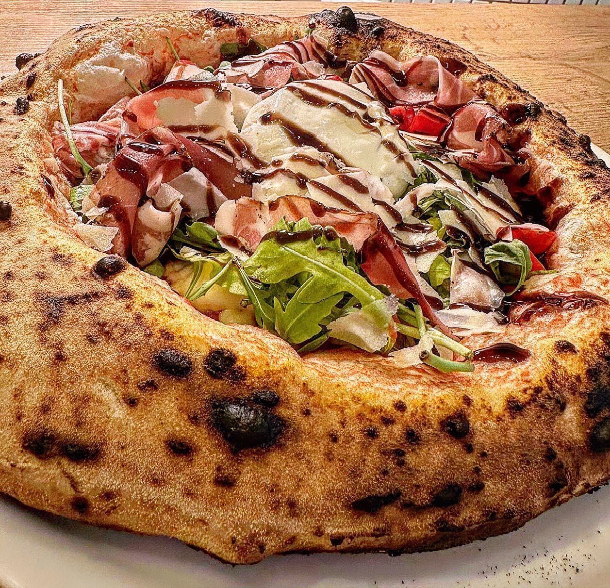 Gros plan sur une pizza à la croûte épaisse et grillée. Garnie de prosciutto, de roquette et d'un filet de glaçage balsamique.