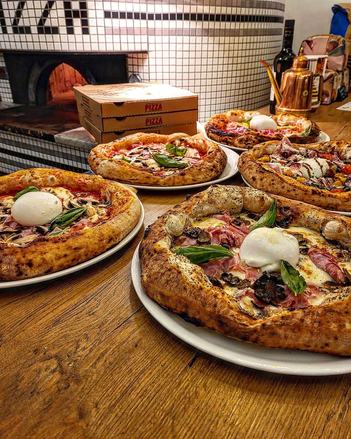 Gros plan sur plusieurs pizzas à la croûte croustillante et aux garnitures variées, disposées sur une table en bois ; le four est visible en arrière-plan.