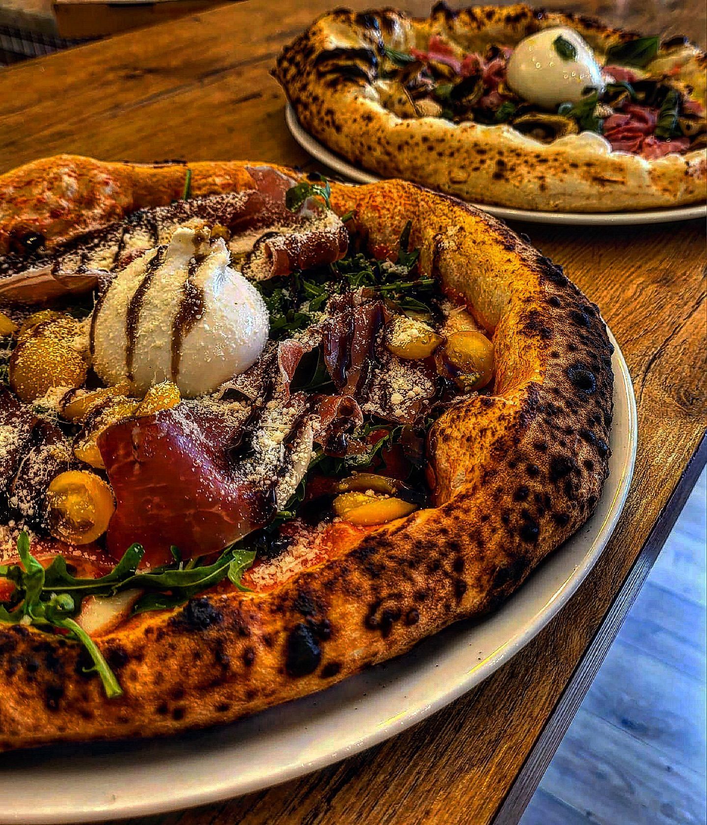 Deux pizzas sur des assiettes, à la croûte grillée et garnies de diverses garnitures : prosciutto, burrata, roquette et maïs.