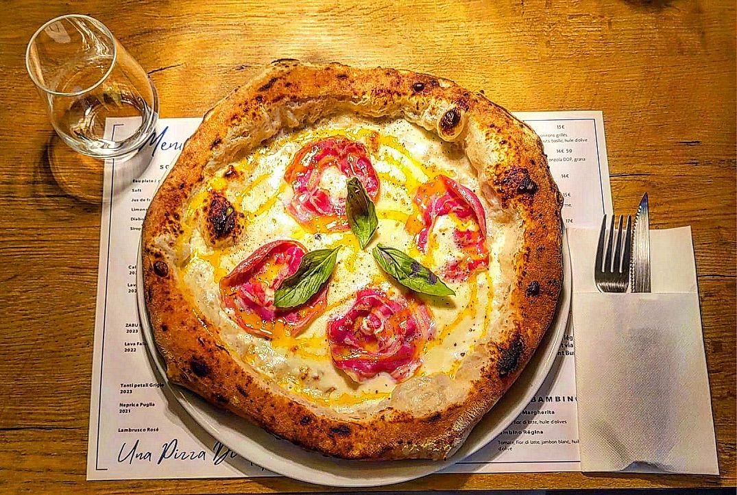 Pizza à pâte épaisse, garnie de sauce blanche, de viande rosée et de feuilles de basilic, sur une table en bois.