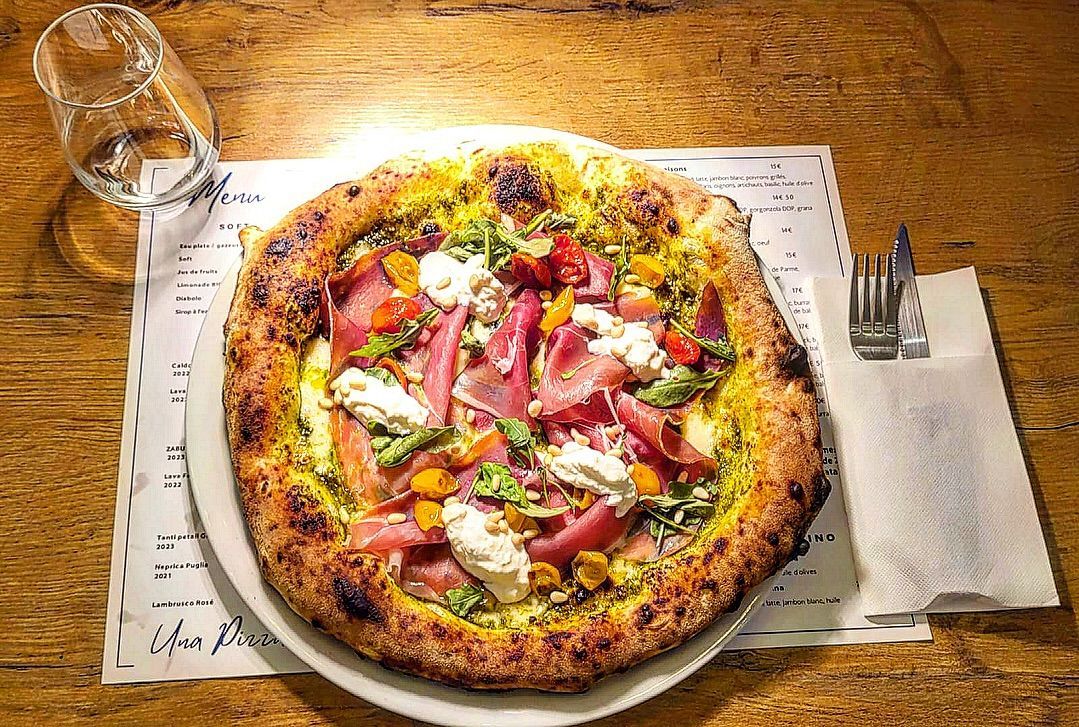 Pizza garnie de prosciutto, de fromage et de verdure, servie sur une assiette blanche avec des couverts, un menu et un verre, le tout sur une table en bois.