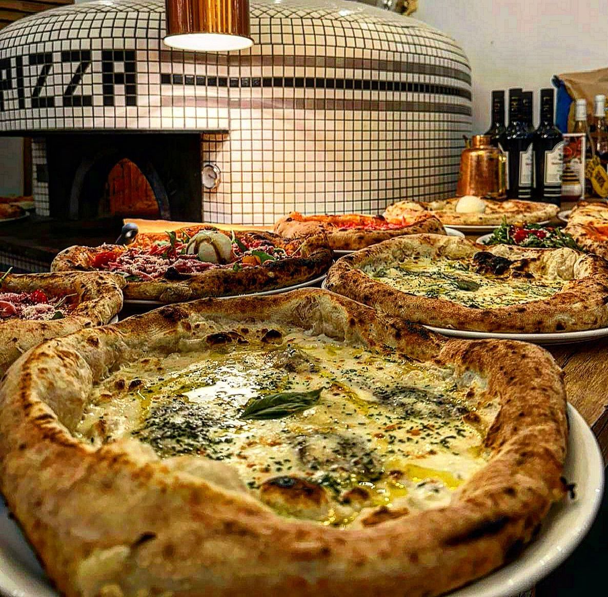 Des pizzas exposées devant un four à briques sur lequel est inscrit PIZZA ; des bouteilles de vin en arrière-plan.