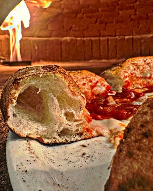 Gros plan sur une pizza cuite au feu de bois avec une croûte croustillante, de la sauce visible et du fromage fondu ; le feu du four en briques est visible en arrière-plan.