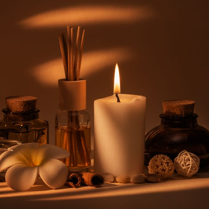 Une bougie blanche allumée, un diffuseur de parfum, une fleur blanche et de petits objets de décoration disposés sur une surface aux tons chauds.
