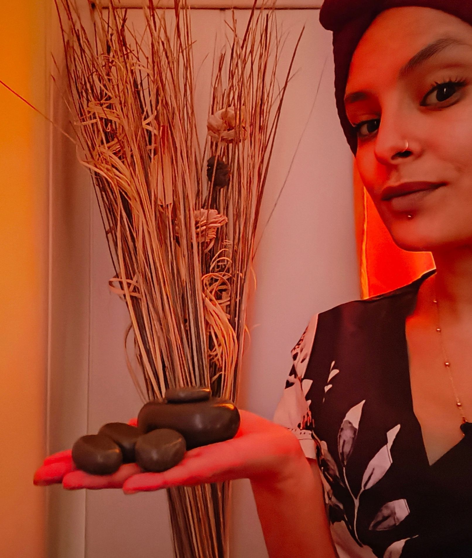 Une personne tient quatre pierres de massage noires et lisses dans sa paume, avec des brindilles séchées décoratives en arrière-plan.