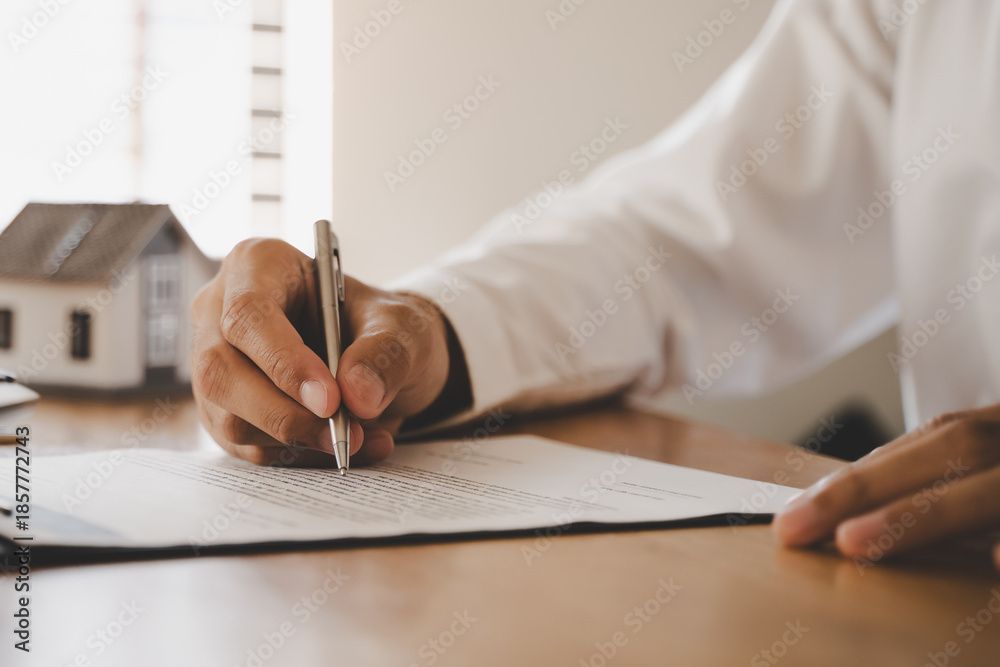 Eine Person unterzeichnet ein Dokument an einem Schreibtisch; im Hintergrund ist ein kleines Hausmodell zu sehen.