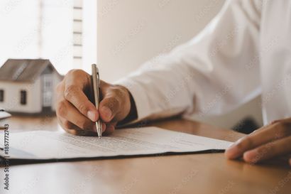 Eine Person unterzeichnet ein Dokument an einem Schreibtisch; im Hintergrund ist ein kleines Hausmodell zu sehen.