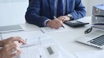 Eine Person im Anzug sitzt an einem Schreibtisch und liest Dokumente; ein Taschenrechner und ein Laptop sind vorhanden.