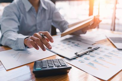 Eine Person prüft Finanzdokumente mithilfe eines Taschenrechners, Diagrammen und eines Stifts auf einem Schreibtisch.