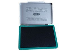 Almofada de tinta Pelikan aberta com superfície de tinta escura e borda verde. A tampa é prateada com etiqueta da marca.