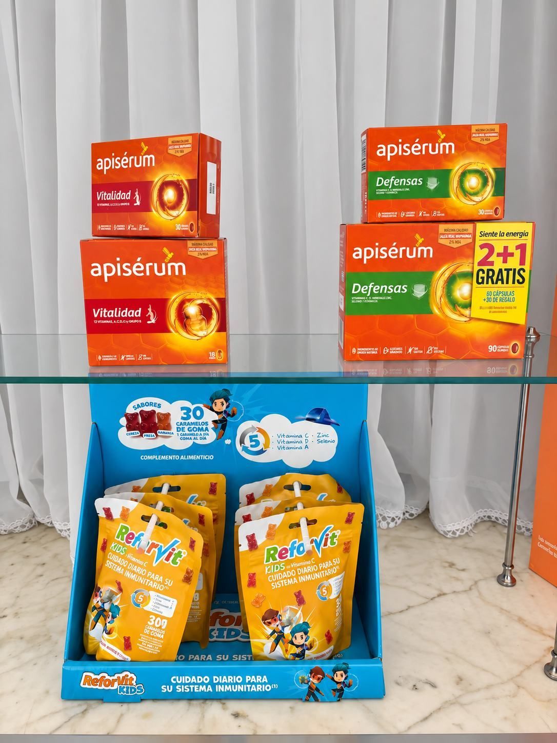 Exhibición colorida de productos apilados sobre una mesa de cristal frente a unas cortinas blancas, con artículos envasados de color naranja y amarillo.