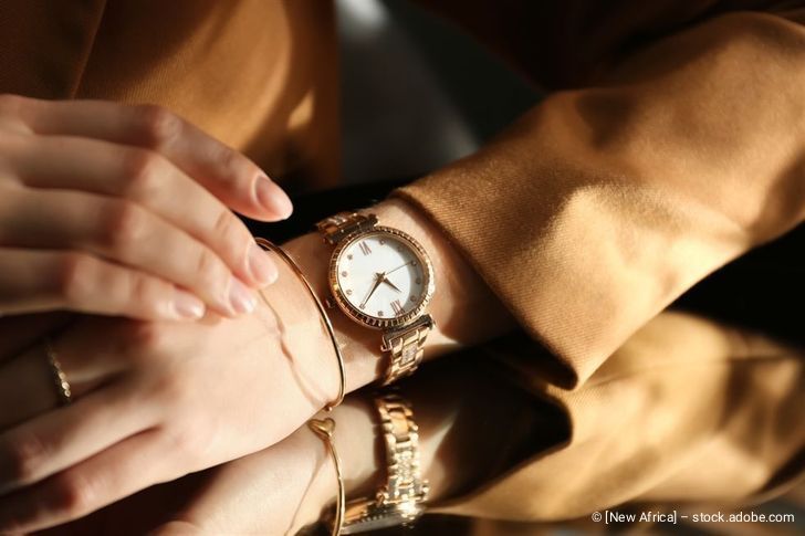Die Hand einer Frau mit einer goldenen Uhr und Armbändern ruht auf dem Ärmel einer braunen Jacke.
