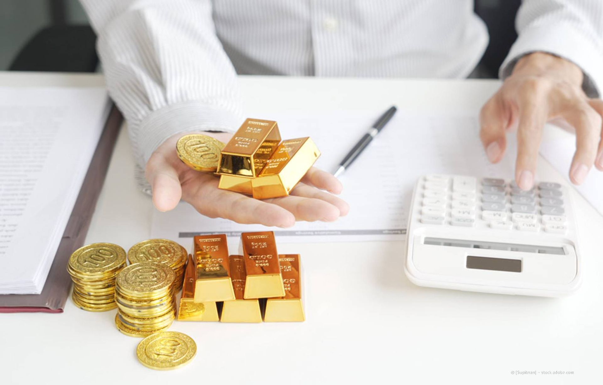 Eine Person hält Goldbarren und Münzen in den Händen und benutzt einen Taschenrechner auf einem Schreibtisch mit Papierkram.