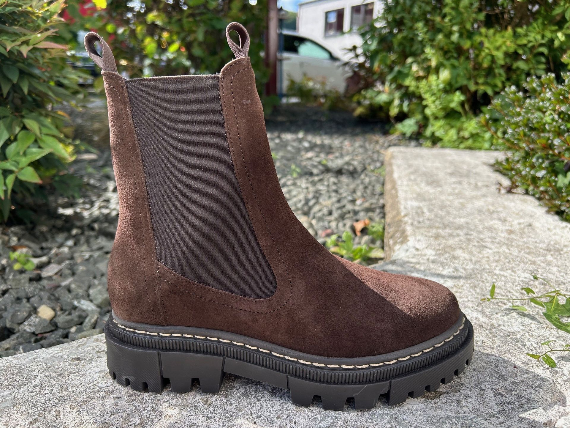 Chelsea-Stiefel aus braunem Wildleder mit dicker schwarzer Sohle, die auf einer Steinoberfläche ruht.