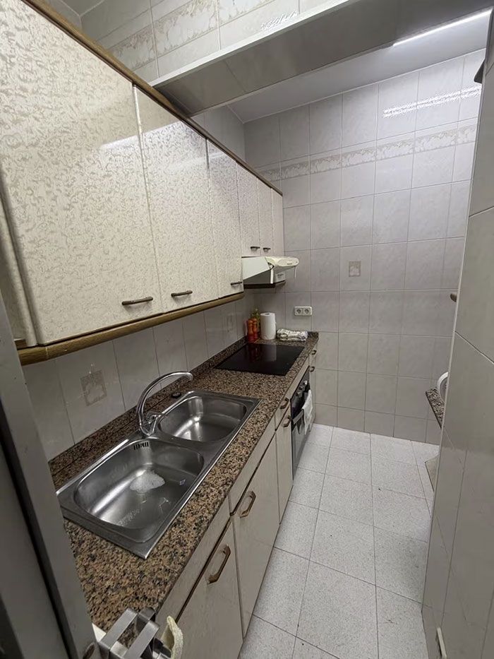 Cocina estrecha con encimera de granito, fregadero doble, armarios y paredes y suelo de azulejos blancos.