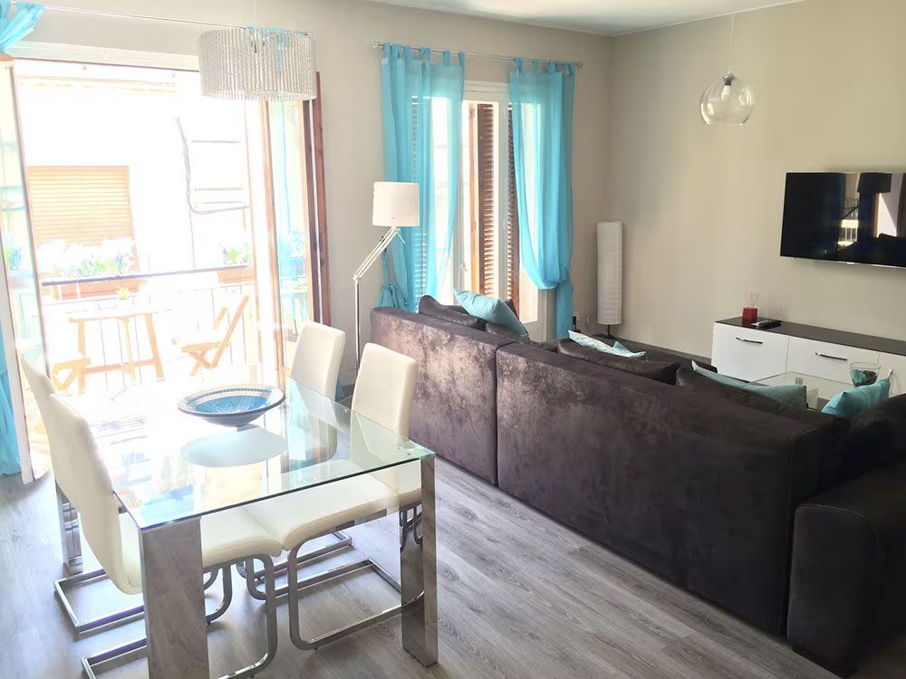 Luminoso salón-comedor con mesa de cristal, sofá oscuro, televisor de pared y cortinas azules cerca de una puerta que da al balcón.