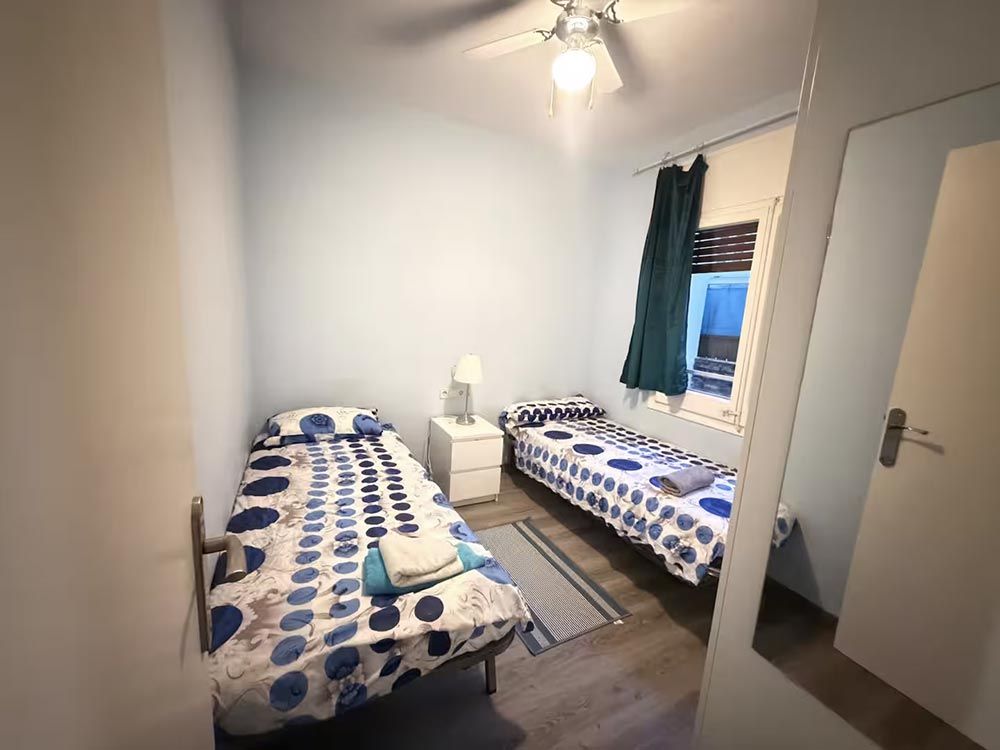 Habitación pequeña con dos camas individuales, ropa de cama con estampado azul, una mesita de noche, ventana y puerta abierta.