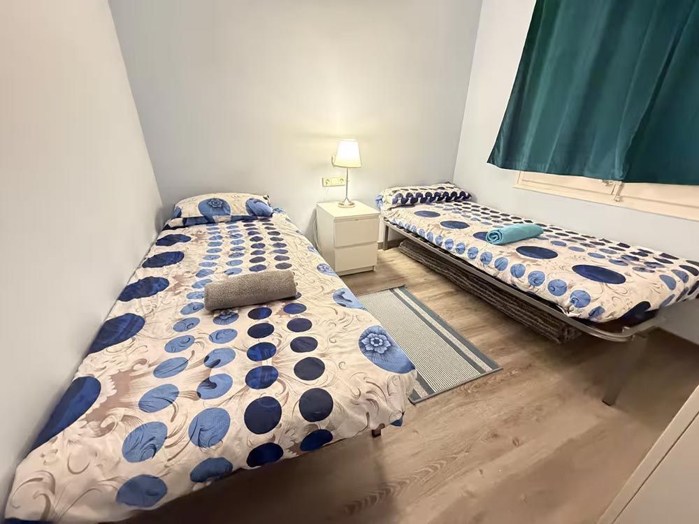Habitación pequeña con dos camas individuales, ropa de cama azul de lunares, una lámpara y una ventana con cortinas oscuras.