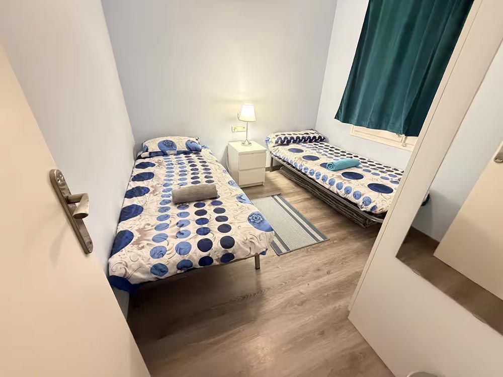 Habitación pequeña con dos camas individuales, ropa de cama con estampado azul, lámpara de noche y armario con espejo.